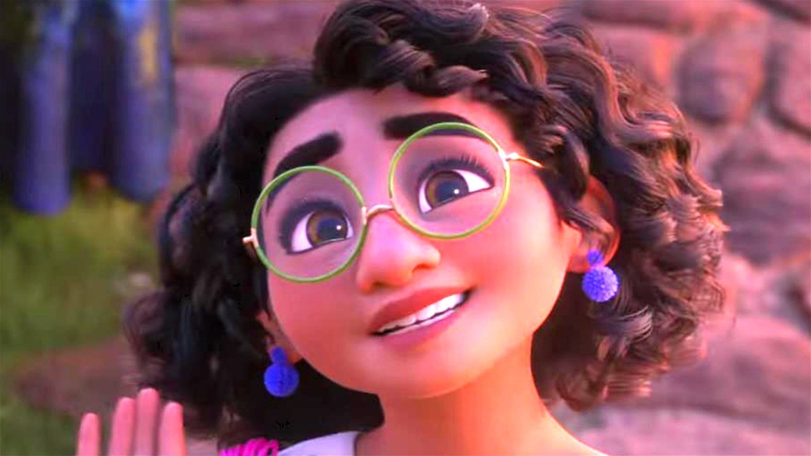 Image of Mirabel from the Disney film 'Encanto.' She is a brown-skinned girl with curly, dark hair, thick, dark eyebrows, green, thin-rimmed glasses and purple earrings. She's waving and smiling nervously in this close-up. 