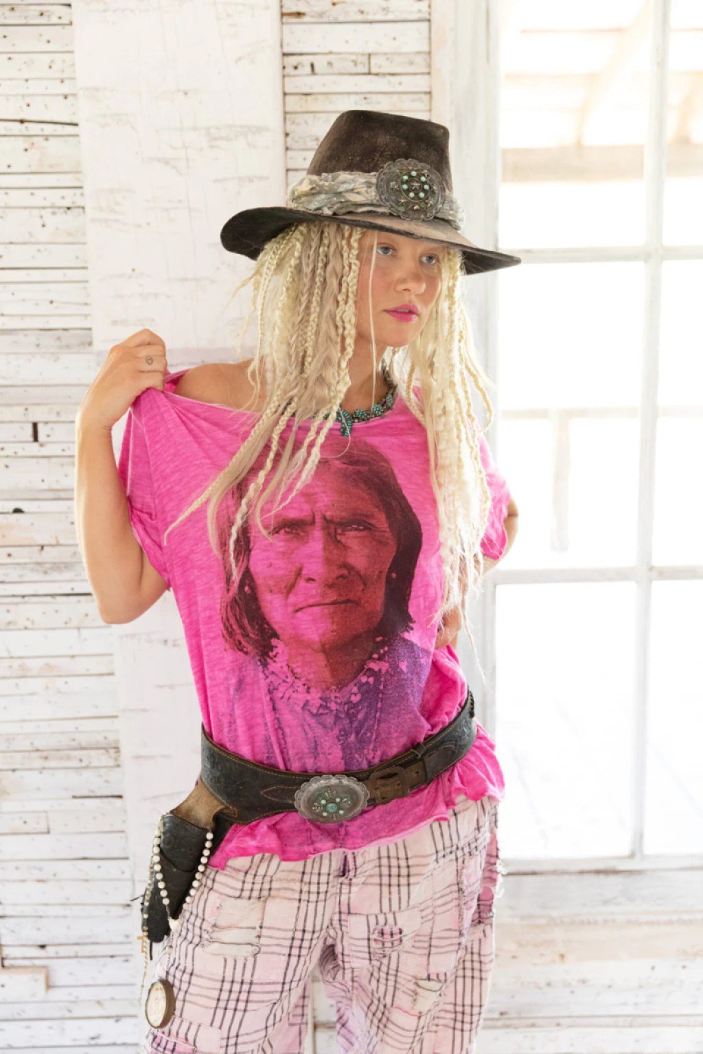 A blonde woman with lots of thin, messy braids and a brown cowboy hat wear a pink t-shirt with a black and white picture of Goyaałé on it.
