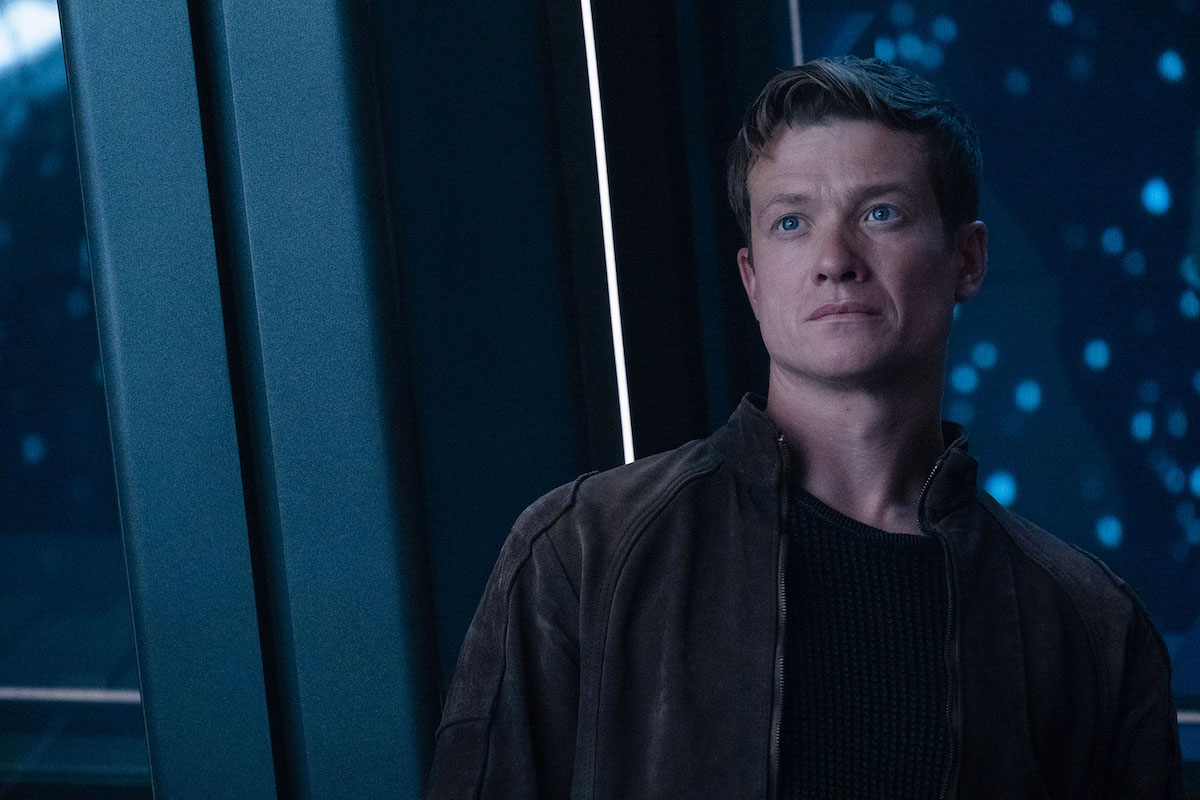Jack Crusher stands against a window in a spaceship, looking concerned.