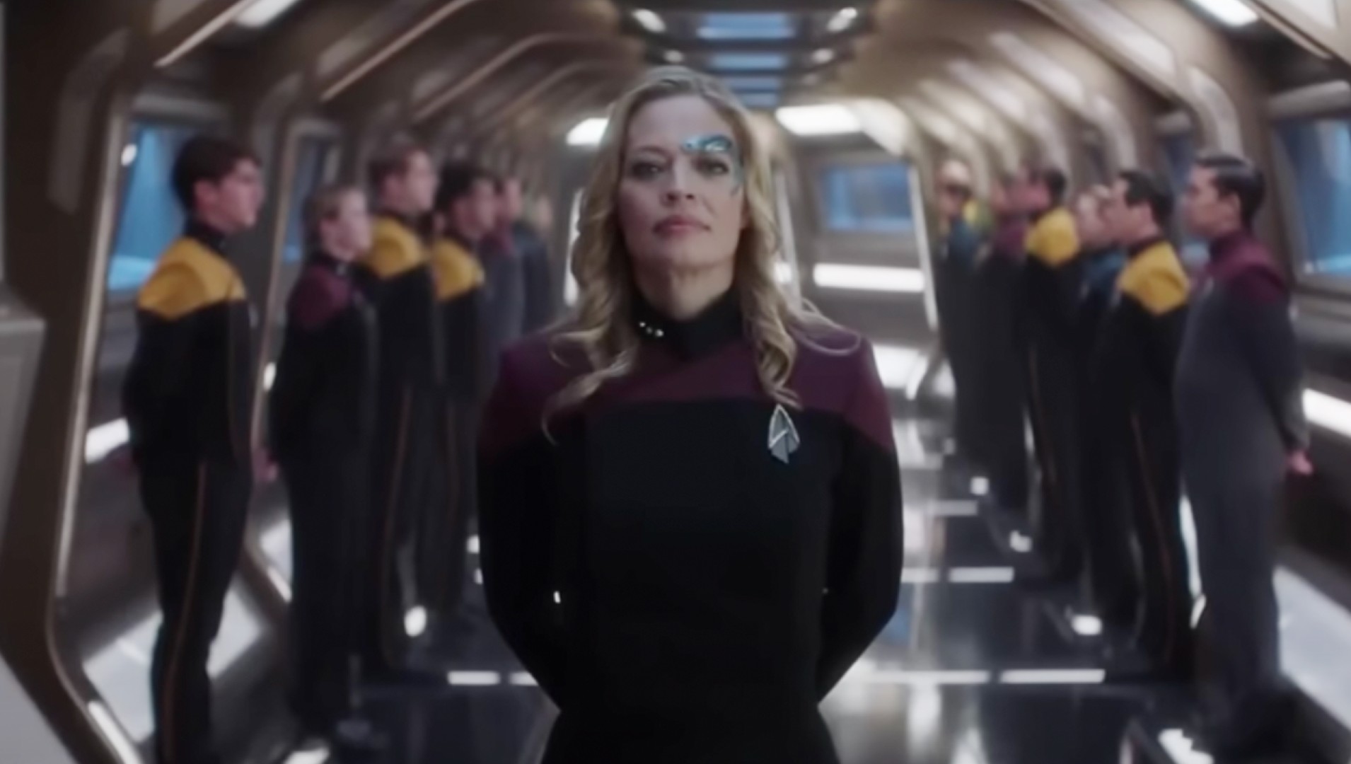 Jeri Ryan as Seven of Nine in a scene from 'Star Trek: Picard' on Paramount Plus. She is standing At Ease in a ship corridor. Behind her are two rows of Starfleet officers on either side