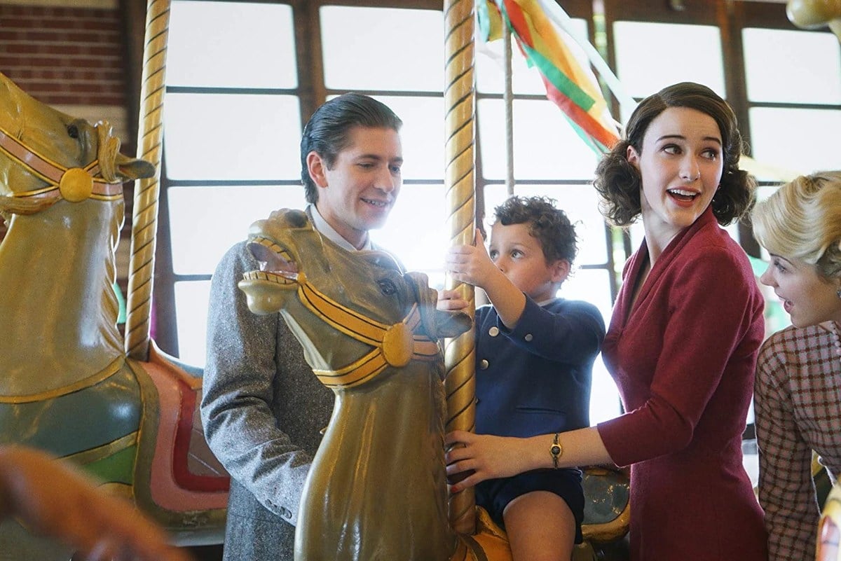Michael Zegen as Joel, Matteo Pascale as Ethan, and Rachel Brosnahan as Midge in Amazon's 'The Marvelous Mrs. Maisel.' This is a 1950s/60s period piece. They're standing on a merry-go-round and Ethan is on a horse as his parents stand on either side. Joel is a white, Jewish man with slicked back hair wearing a grey suit. Ethan is a white toddler with curly, dark hair wearing a navy blue suit jacket and shorts. Midge is a white, Jewish woman with chin-length, curled hair wearing a red dress with long sleeves. 