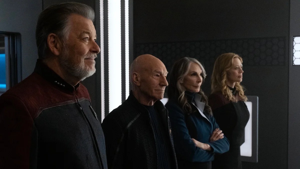 Jonathan Frakes as Riker, Patrick Stewart as Picard, Gates McFadden as Dr. Crusher, and Jeri Ryan as Seven of Nine in a scene from 'Star Trek: Picard' on Paramount Plus. They are standing in a row smiling at something in front of them. Riker is a white man with salt-and-pepper hair and a beard wearing a red and black Starfleet uniform with Captain pips visible on his collar. Picard is a white, bald man wearing a black jacket and black shirt. Crusher is a white woman with dark hair that has thick, white streaks in the front. She's wearing a blue jacket and her arms are folded. Seven is a white woman with long, blonde hair dressed all in black. 