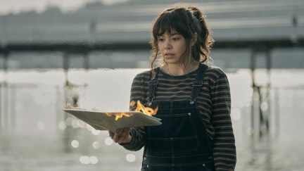 Jos stares at a burning piece of paper in her hand, with the ocean behind her.