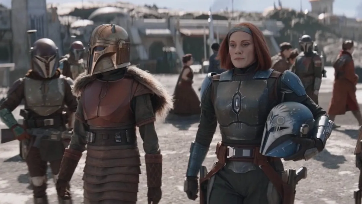 Emily Swallow as The Armorer and Katee Sackhoff as Bo-Katan in 'The Mandalorian' on Disney+. They are walking through a busy area. The Armorer is in full Mandalorian armor, while Bo-Katan is in her armor, but has her helmet off and in one arm. Her face, grey headband, and chin-length red hair are visible. 