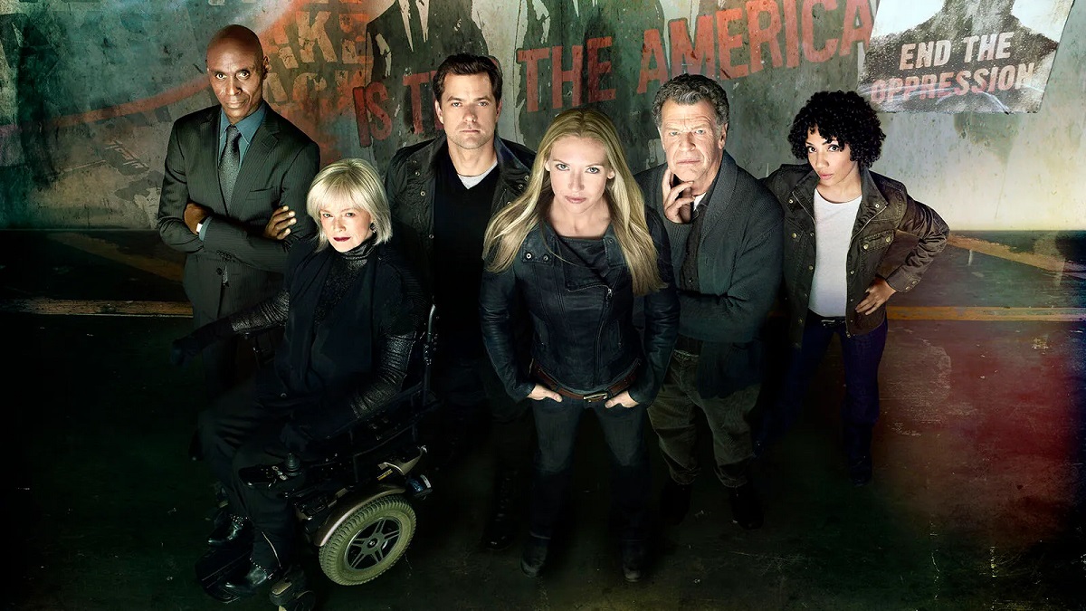 Lance Reddick, Blair Brown, Joshua Jackson, Anna Torv, John Noble, and Jasika Nicole in character in a promotional image for the FOX show, 'Fringe.' Reddick is a bald, Black man in a black suit with a blue buttondown and a black tie standing with his arms folded. Blair is a white woman with white chin-length hair dressed all in black and sitting in a wheelchair. Jackson is a white man with short dark hair standing behind Brown and Torv in a black jacket, black pullover with a white t-shirt underneath. Torv is in the center of the photo standing with her hands in her pockets. She's a white woman with long blonde hair dressed all in black. Noble stands behind Torv. He's a white man with salt-and-pepper wavy hair wearing a grey cardigan over a dark buttondown and dark pants. He has one hand on his face. Nicole stands behind Noble. She is a light-skinned Black woman with curly black hair that comes down to her ears. She wears a black leather jacket over a white t-shirt and dark pants. Behind them is a wall with torn protest posters on it. One reads "End the Oppression."
