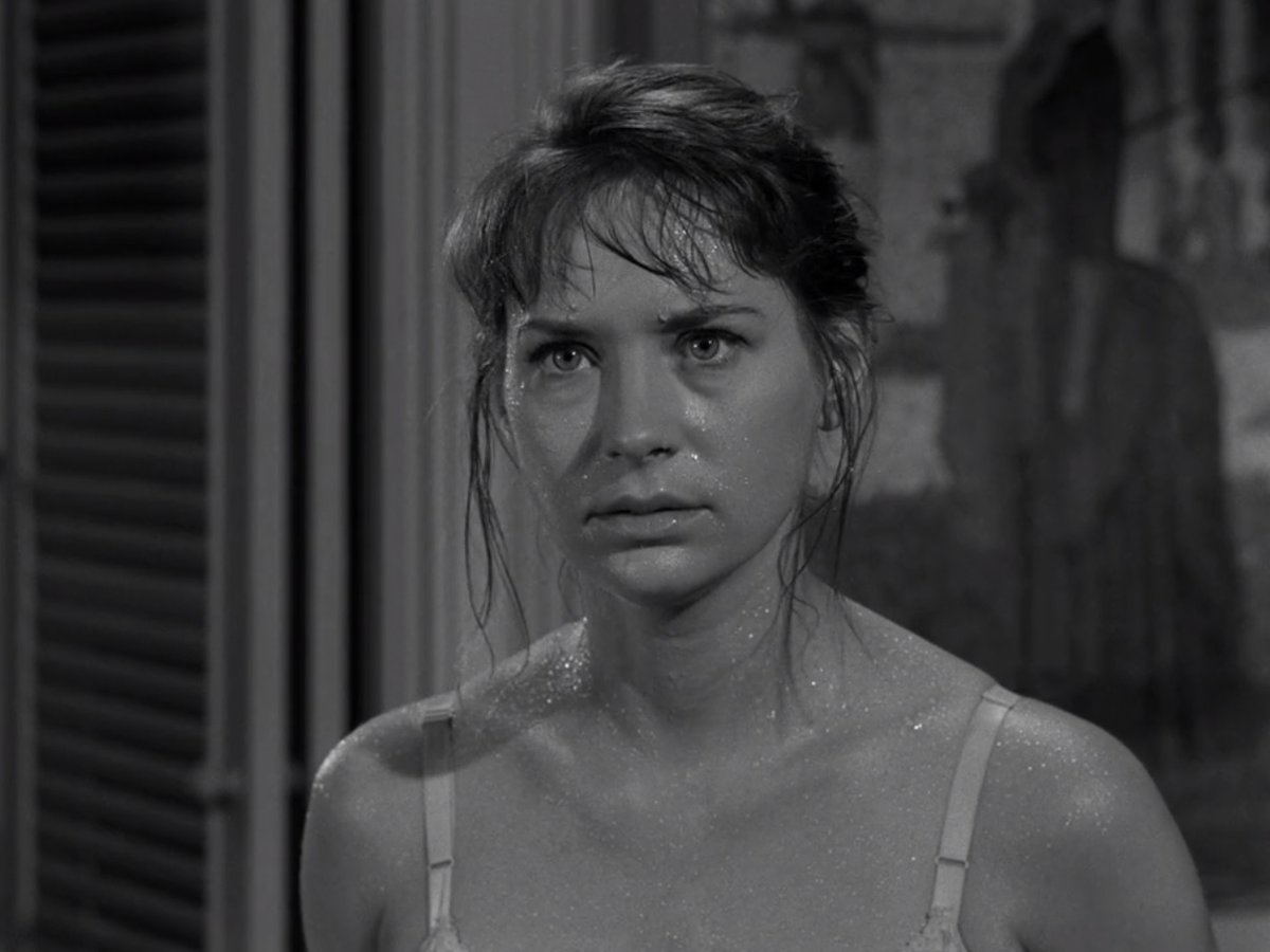 Lois Nettleton as Norma in the "Midnight Sun" episode of 'The Twilight Zone.' It's a black and white image of Norma from the shoulders up. She's a white woman standing in her living room, her long, brown hair is up in a bun, and loose tendrils hang down on either side of her face and she has bangs. She is dripping with sweat, and we can see the straps of the slip she's wearing on her shoulders.