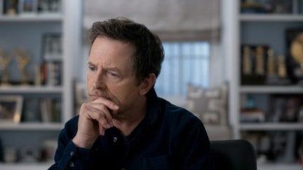 Michael J. Fox rests his chin on his hand contemplatively in front of a window and bookshelves.