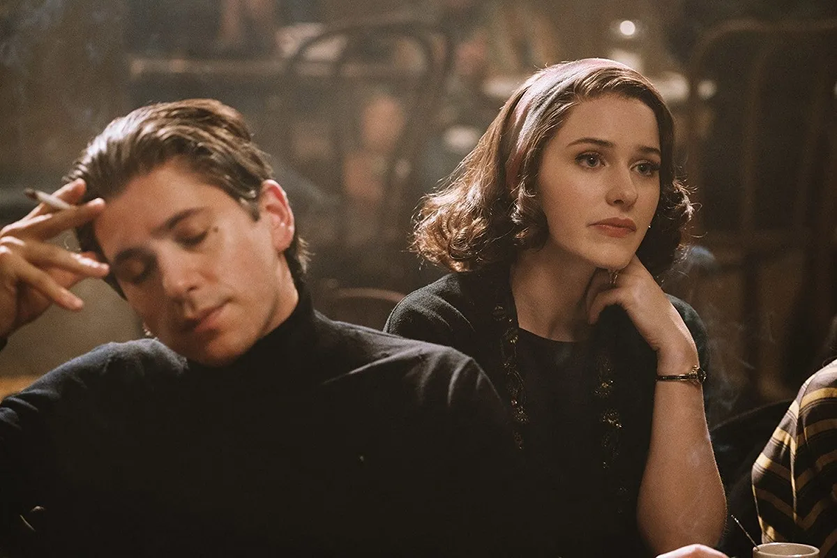 Michael Zegen as Joel, and Rachel Brosnahan as Midge in Amazon's 'The Marvelous Mrs. Maisel.' This is a 1950s/60s period piece. They're sitting at a table in a comedy club. Joel is a white, Jewish man with slicked back hair wearing a black turtleneck and holding a lit cigarette. He's holding his head in his cigarette hand as he sits with his eyes closed, frustrated with something. Midge is a white, Jewish woman with chin-length, curled hair with a scarf tied in it wearing a black shirt with three-quarter sleeves, a chunky, brown necklace, and a gold bracelet. Her elbow is on the table as she props her chin up with her hand and looks out into the distance, disappointed.