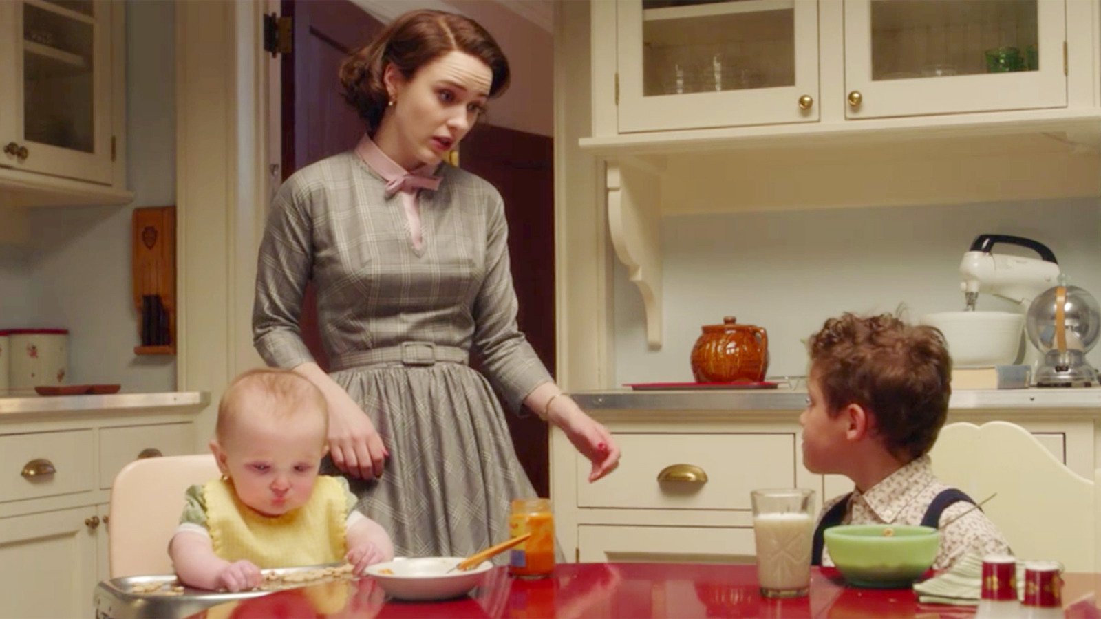 Rachel Brosnahan as Miriam "Midge" Maisel with baby Esther and older son Ethan in a scene from Amazon's 'The Marvelous Mrs. Maisel.' The kids are seated at the kitchen table as Midge stands talking to Ethan. She is a white woman with bobbed reddish-brownish hair and wearing a grey, belted, 1950's style dress with a pink collar and bow at her neck. 