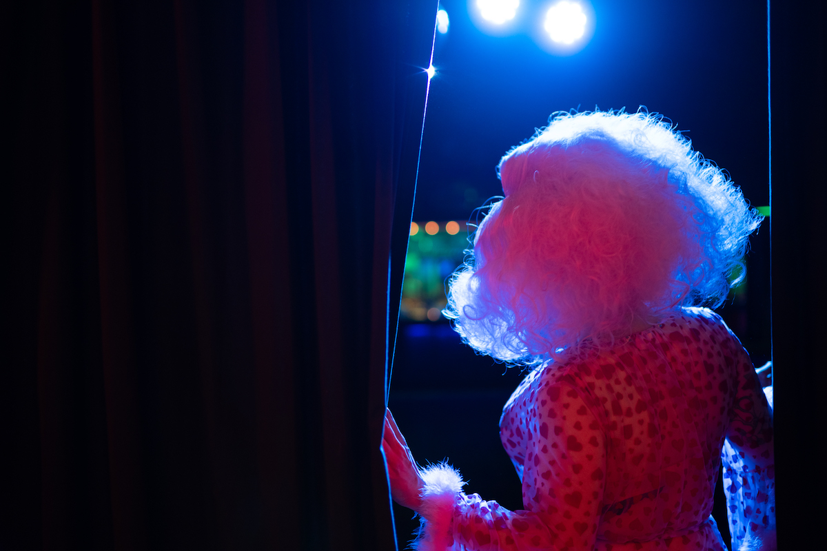 A drag queen is seen from behind, moving a stage curtain aside to look at the audience.