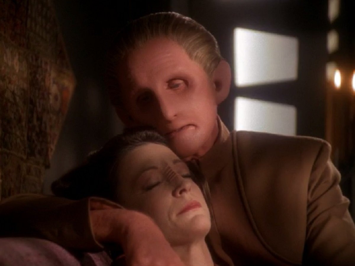 Rene Auberjonois as Odo and Nana Visitor as Kira Nerys in 'Star Trek: Deep Space Nine.' Odo is a white Changeling who's adopted a male humanoid shape with blond, slicked back hair and wearing a brown Starfleet uniform. He is holding Nerys, who is a white Bajoran woman with ridges along the bridge of her nose. Her eyes are closed. 