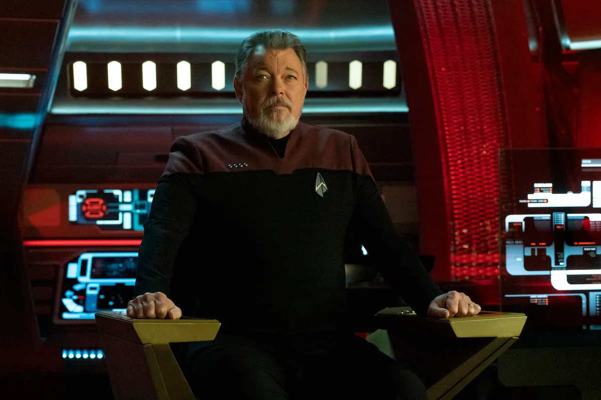 Captain Riker in Picard: An older white man with grey hair and beard sits in the captain's chair, wearing a red and black Starfleet uniform