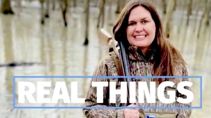 Sarah Huckabee Sanders awkwardly smiling as she stands in the woods wearing camo gear and holding a rifle.