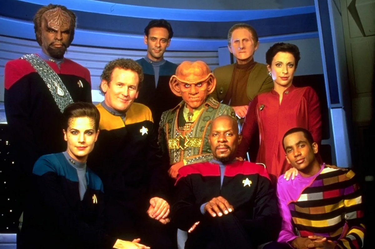 Promotional image of the cast of 'Star Trek: Deep Space Nine.' There are nine actors in the photo, all in character and on the set. Five are made up as alien characters, the rest are human. Two women, one boy, and the rest are men.