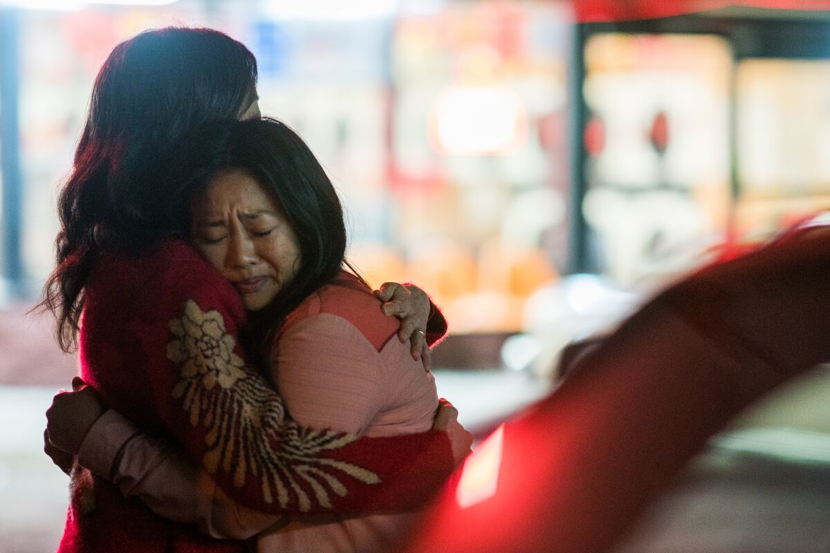 Michelle Yeoh as Evelyn and Stephanie Hsu as Joy in 'Everything Everywhere All at Once' from A24. As they stand in a parking lot, they are in focus while the storefronts and cars around them are out of focus. They are hugging. We see Evelyn from the side, but not her face. She has long, black hair and is wearing a long-sleeved, red sweater with a gold design on the sleeves. Joy is a young Chinese woman who is pressing her head against Evelyn's shoulder as they embrace. Her eyes are closed, and she has a pained expression on her face. She's wearing a pink jacket. 