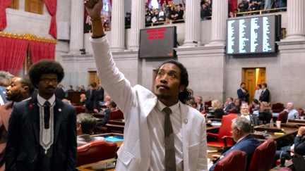 Democratic Rep. Justin Jones wears a white suit and raises his fist in the air, looking up towards protesters above him in the State Capitol.