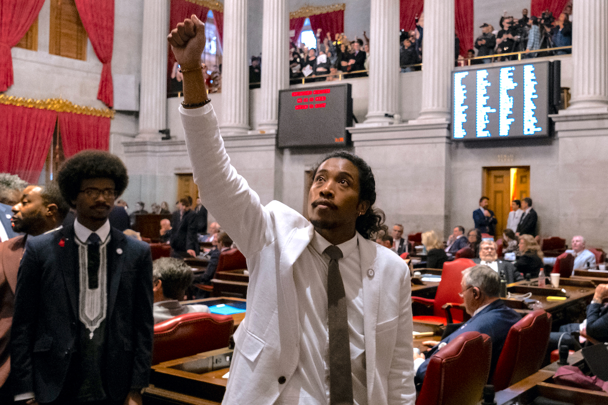 Democratic Rep. Justin Jones wears a white suit and raises his fist in the air, looking up towards protesters above him in the State Capitol.