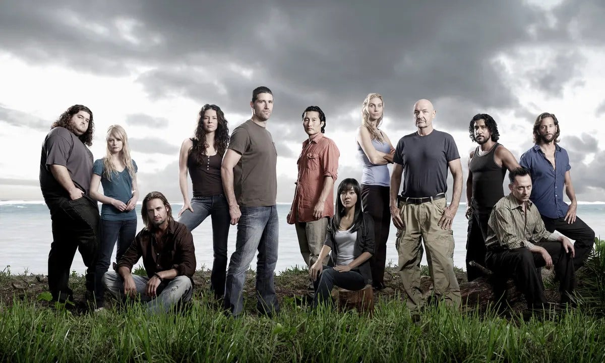 Promotional image of the cast of the ABC show 'lost.' There are 12 actors in the photo, and they're all in character outdoors sitting or standing together in the grass overlooking the ocean. Three are seated, everyone else stands around them. Four women, the rest men. The sky behind them has dark clouds.