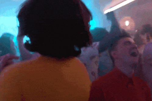 GIF of James Lance as Trent and Billy Hughes as Colin on Apple TV+'s 'Ted Lasso.' Trent is an older, white man with longish brown hair with white streaks in it and wearing a yellow t-shirt. Colin is a younger white man with short dark hair in an orange buttondown shirt. Thy are both dancing in a crowd at a gay bar.