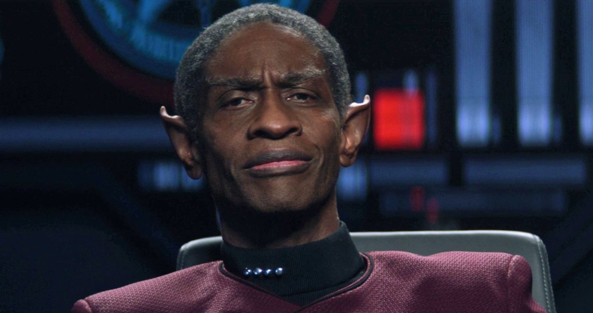 Tim Russ as Tuvok in an episode of 'Star Trek: Picard' on Paramount+. He is a Black Vulcan sitting in the captain's chair on a ship with salt-and-pepper, close-cropped hair, pointy ears, and is wearing a red Starfleet uniform with the rank of Captain. He's smiling a soft smile, which is suspicious for a Vulcan.