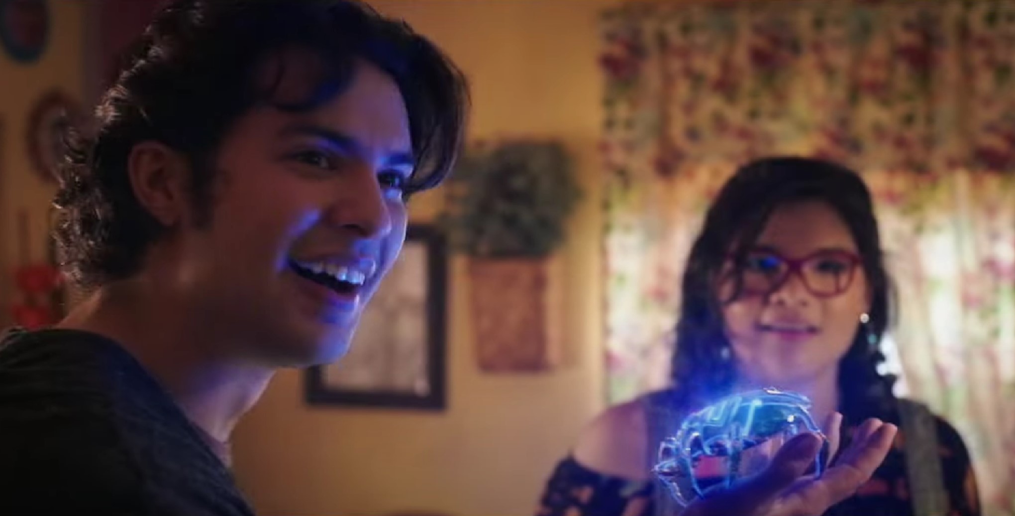 Screenshot of Xola Maridueña as Jaime Reyes and Belissa Escobedo as Milagro Reyes in the DCEU's 'Blue Beetle.' They are standing in a livingroom with floral curtains. Jaime is a Mexican man with brown skin and shaggy dark hair, wearing a black shirt. He's turned to someone and smiling as he holds a glowing, blue scarab in his hand. Milagro stands in the background. She's a Mexican girl with shoulder-length dark hair, glasses, and is wearing an off-the-shoulder black shirt. She's looking at the scarab in amazement. 