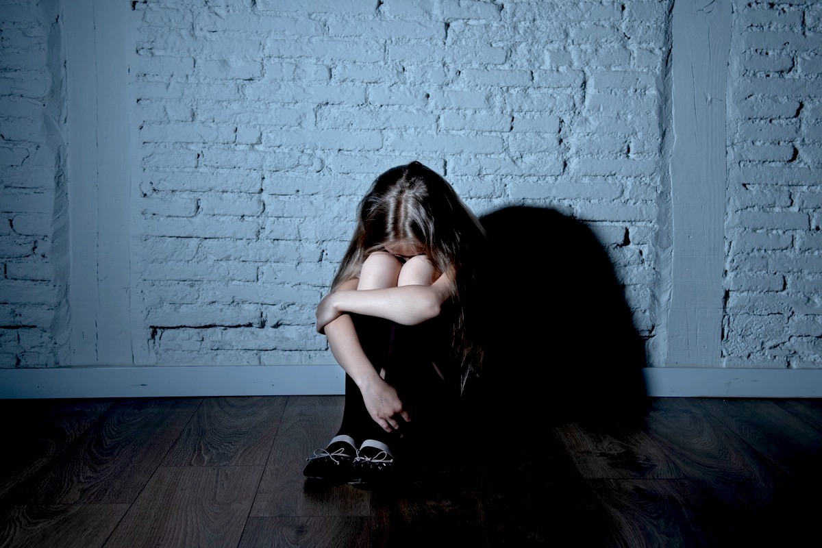 A young girl, sitting on the floor, with her knees pulled up to hide her crying face.
