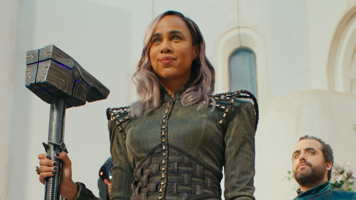 Zawe Ashton, dressed in a grey uniform, smiles as she holds a giant hammer.