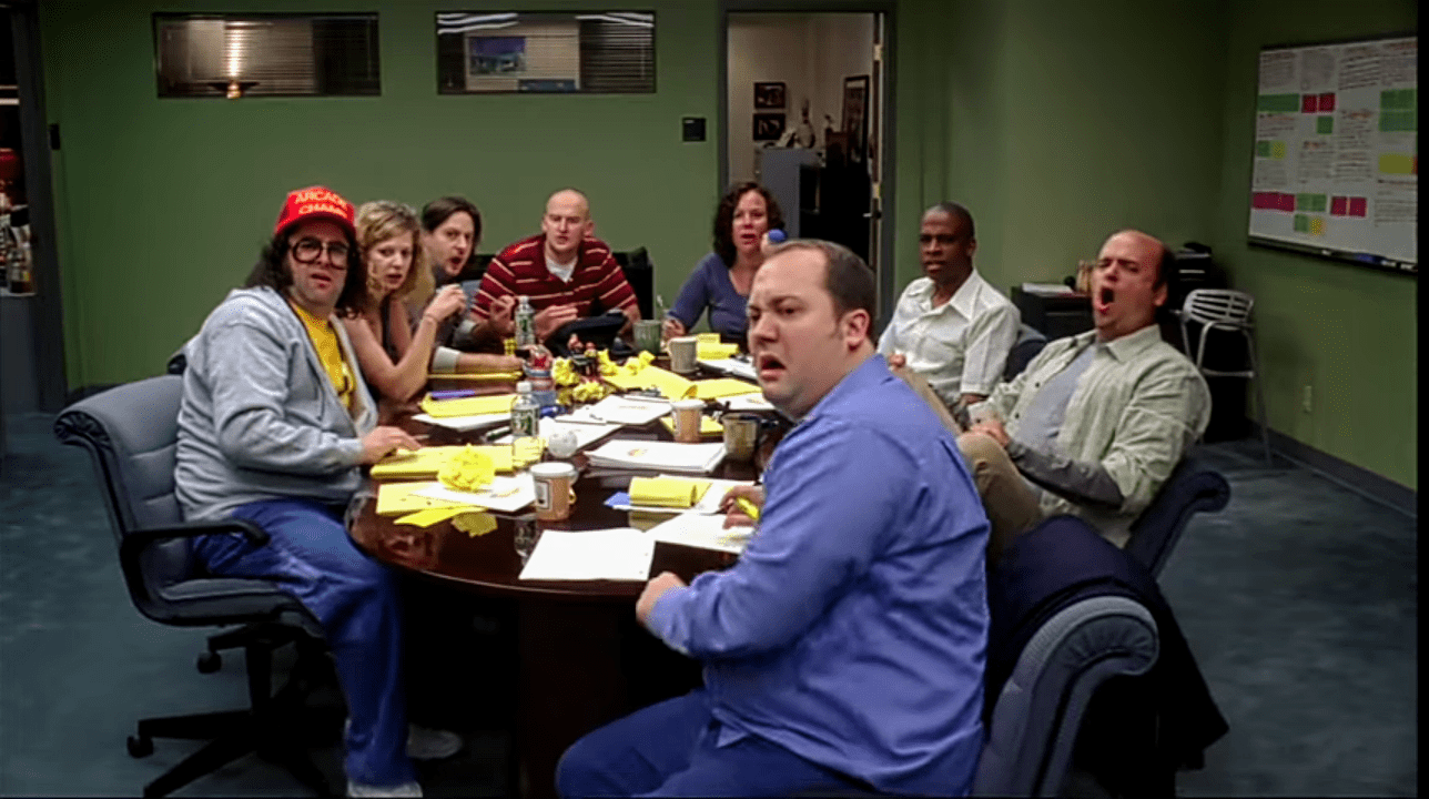 Image of the fictional writers' room on NBC's '30 Rock.' Eight writers sit around a cluttered conference table all looking at something off camera in shock. There are six men and two women. Everyone is white except for "Toofer," played by Keith Powell, who is Black. 