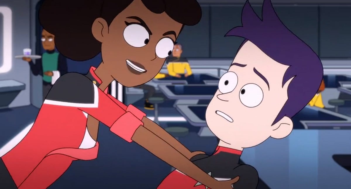 Image of Mariner and Boimler in a scene from the animated series, 'Star Trek: Lower Decks.' Mariner is a Black woman  in a red Starfleet uniform with her hair in a ponytail, and Boimler is a white man with purple hair also in a Starfleet uniform. Mariner is leaning over him with a menacing smile on her face. He looks concerned.