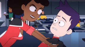 Image of Mariner and Boimler in a scene from the animated series, 'Star Trek: Lower Decks.' Mariner is a Black woman in a red Starfleet uniform with her hair in a ponytail, and Boimler is a white man with purple hair also in a Starfleet uniform. Mariner is leaning over him with a menacing smile on her face. He looks concerned.