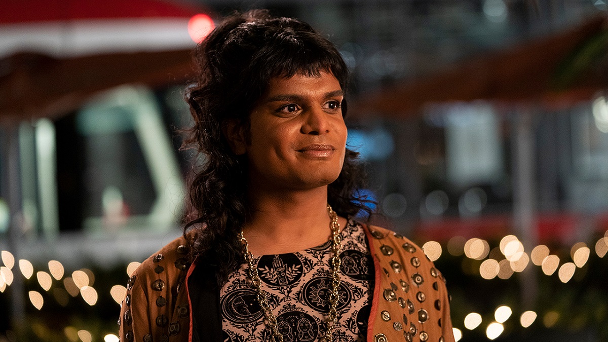 Image of Bilal Baig as Sabi in HBO Max's 'Sort Of.' Sabi is a Pakistani, non-binary femme with long, black hair and bangs. They are standing outdoors wearing a brown jacket with gold rivets all over it, and a patterned beige and black shirt and a thick, gold necklace. Behind them, there are twinkling lights.