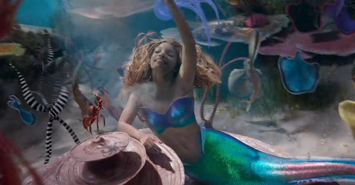 Image of Halle Bailey as Ariel in a scene from 2023's 'The Little Mermaid.' Ariel is a Black mermaid with red hair wearing a purple seashell bra and has a green fish tail. She is smiling with one arm in the air as she leans on some coral where a red crab, Sebastian, is standing. They are "Under the Sea."