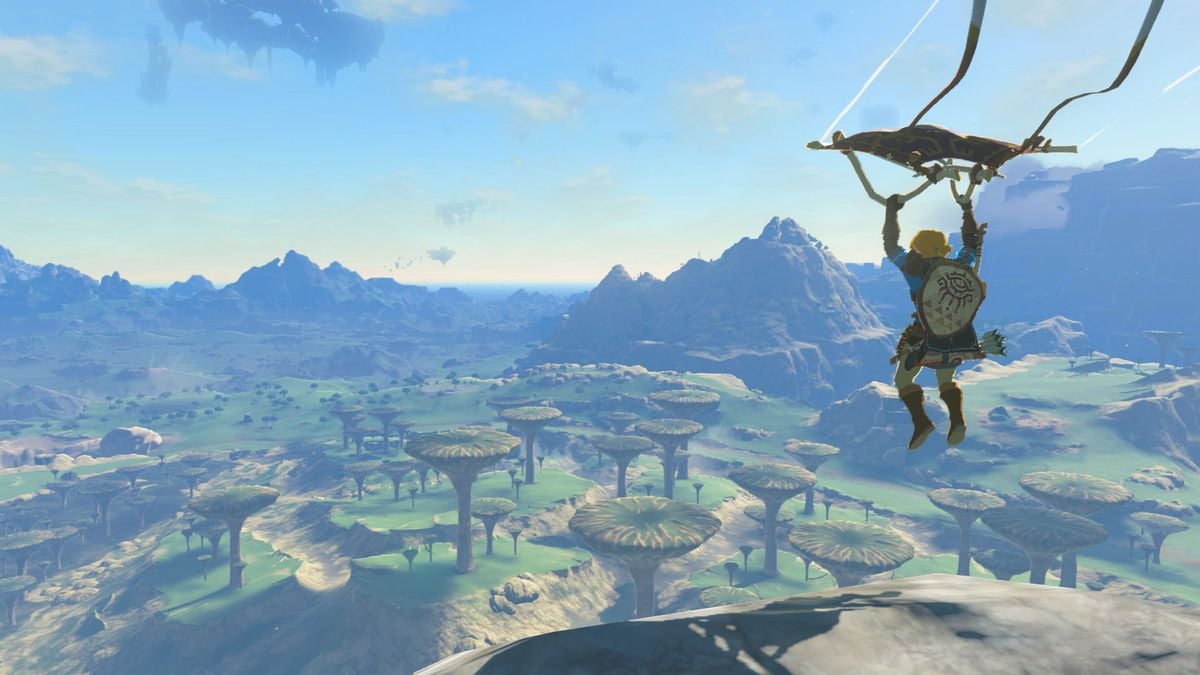 Screenshot of Link in 'The Legend of Zelda: Tears of the Kingdom.' Link, blonde with a large shield on his back and wearing brown boots, is flying through the air holding onto a hang glider. Below him in the sunlight is land full of mushroom-looking platforms and green grass. In the distance are mountains. 