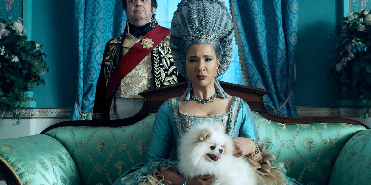 Golda Rosheuvel as Queen Charlotte with a Pomeranian dog on her lap, and Hugh Sachs as Brimsley standing behind her in Queen Charlotte: A Bridgerton Story