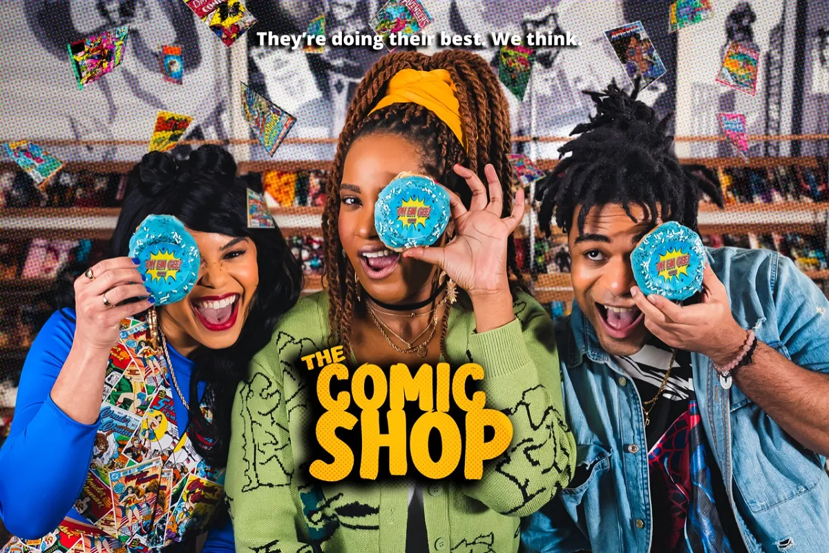 Promotional image for 'The Comic Shop.' The top of the image has the tagline "They're doing their best. We think." The image is of three Black people - two women and one man - each holding a blue frosted cupcake up to one eye that has a logo that reads "Oh Em Gee" on it. Creator Cheyenne Ewulu is in the center. She has long reddish-black braids pulled up in an orange hair wrap and she's wearing a green sweater that buttons down the front. She is flanked by one woman with long dark hair in a long-sleeved blue shirt and a comic book-print vest on her right, and a man with short braids in a blue denim button down over a graphic tee on her left.