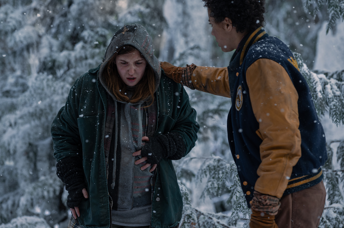 Shauna doubles over in pain while Taissa touches her shoulder. They're in the woods with snow falling.
