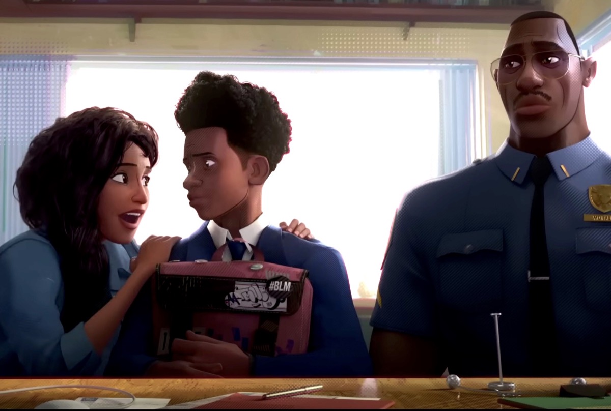 Screenshot from a clip from 'Spider-Man: Across the Spider-Verse.' Miles Morales, an Afro-Latino teenager with an afro and wearing his school uniform of a white buttondown with a tie and a blue blazer as he holds his backpack on this lap, sits between his mother, an Afro-Latina with long, curly dark hair hanging in a loose braid over one shoulder wearing a blue button down, on his right, and his father, a tall, muscular Afro-Latino with close-cropped hair and a thin mustache wearing a police uniform on his left in front of a desk in a high school guidance counselor's office.