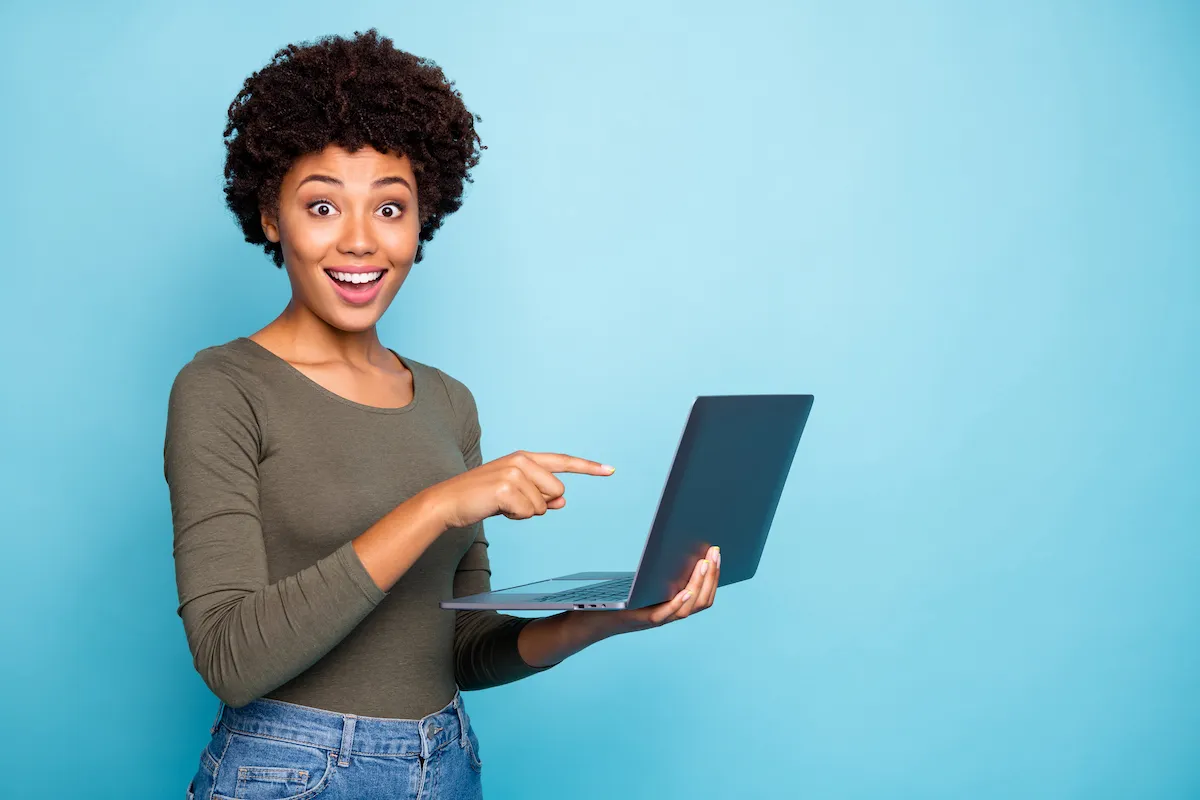 A young Black woman holding her laptop in one hand and pointing at it with the other, with an expression of delight and surprise.