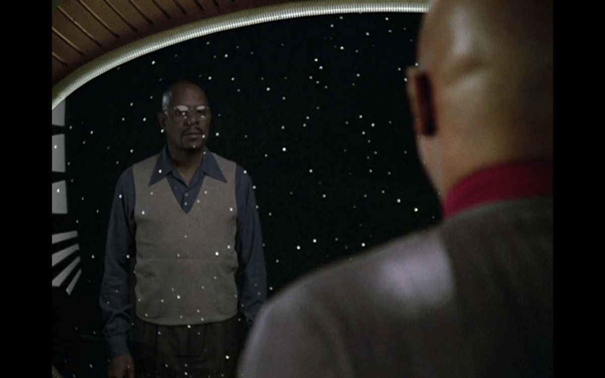 Image of Avery Brooks as Ben Sisko and the reflection of Benny Russell in a scene from the STAR TREK: DS9 episode, 'Far Beyond the Stars.' Brooks is a Black, bald man with a goatee. In the foreground, we see him from behind as he's looking out a window on the space station. He's wearing a grey Starfleet uniform with red at the collar. In the window, we see a reflection of Benny Russell (also played by Brooks) who is wearing glasses and period 1950s menswear - a blue, buttondown shirt with a wide collar under a white pullover vest and dark pants. The vision of Benny is overlaid over the stars in outer space.