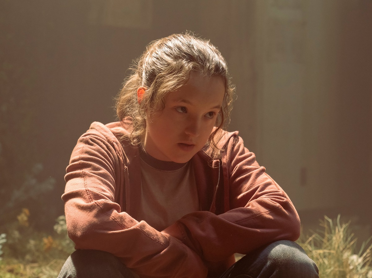 Image of Bella Ramsey as Ellie in a scene from HBO's 'The ast of Us.' Ellie is a white teenage girl with long, brown hair pulled back in a ponytail wearing a red hoodie over a grey shirt and jeans. She's sitting cross-legged in grass with her elbows resting on her knees. She's looking up at someone skeptically. 