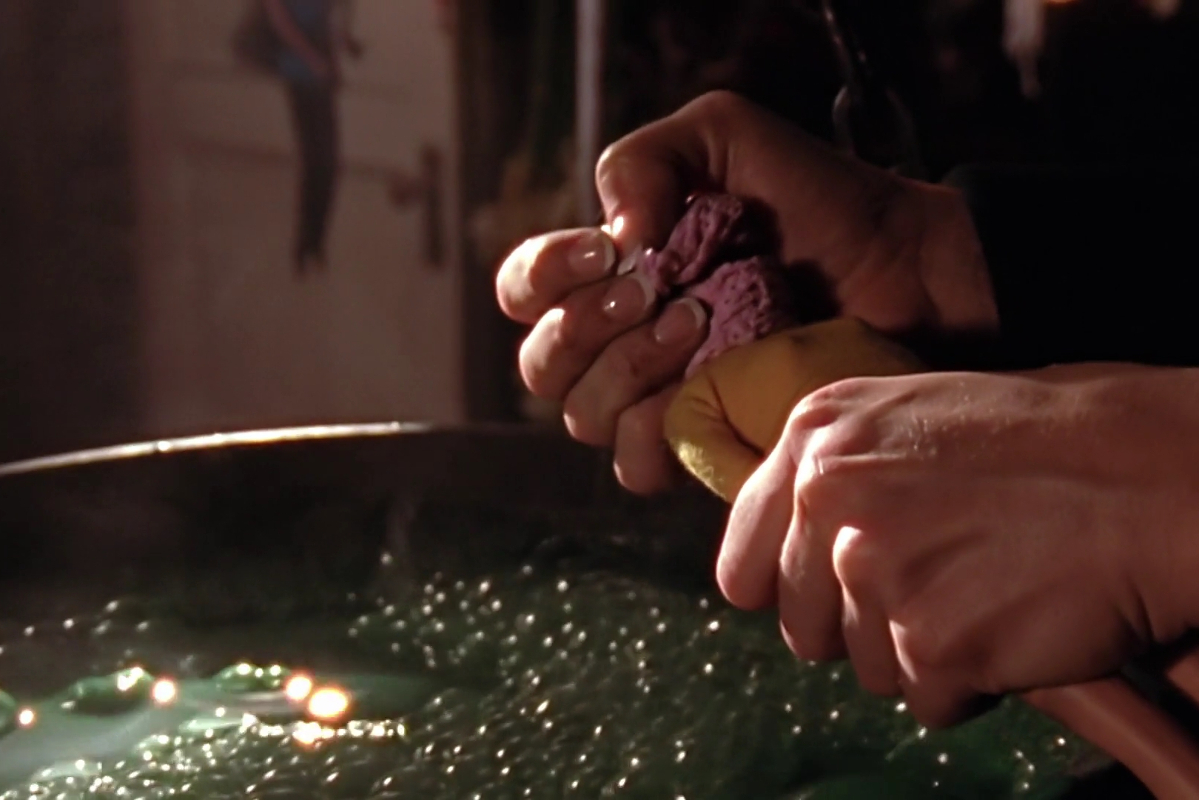 Buffy the Witch episode; a woman's hands lower a cheerleader doll with pink cloth wrapped around it's face into a green, bubbling liquid.