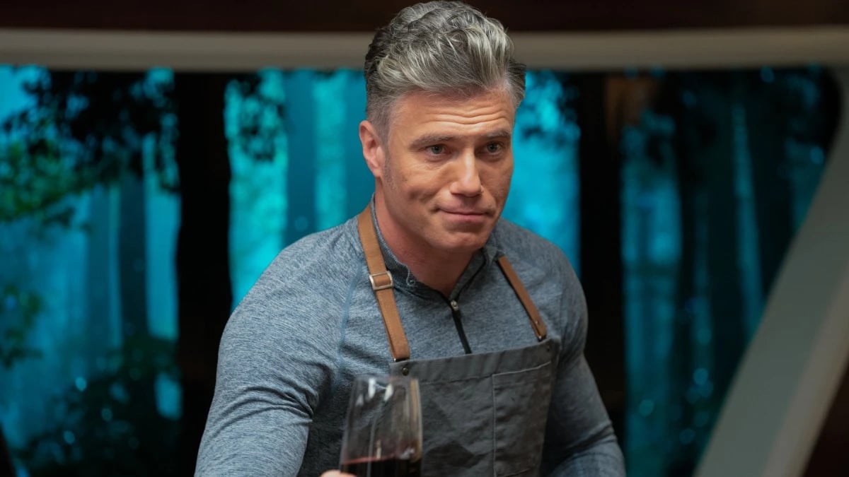 Captain Christopher Pike played by Anson Mount in a scene from 'Strange New Worlds.' He is a white man with mostly silver salt and pepper hair, wearing a blue, long-sleeved cotton zip-up shirt and an apron. He's holding out a glass of red wine with a smirk on his face.