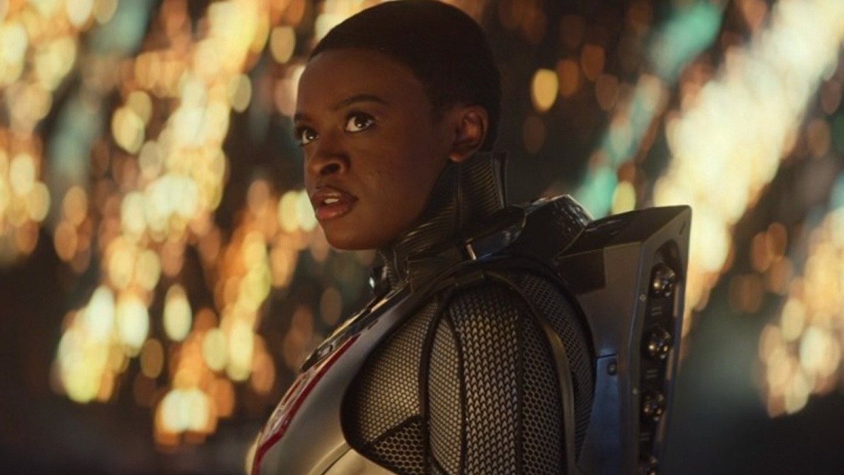 Image of Celia Rose Gooding as Uhura in a scene from the "Children of the Comet" episode of 'Star Trek: Strange New Worlds.' Uhura is a Black woman with close-cropped hair. She's standing to the side with her head turned toward the camera and wearing a grey and black space suit. Behind her are twinkling lights. 