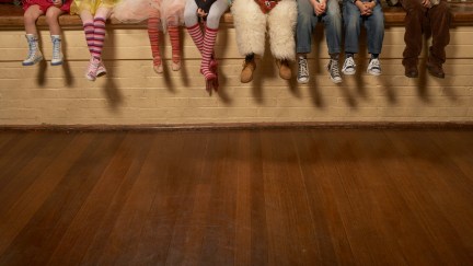 A row of young actors sit on the edge of a stage in various costumes, seen from the waist down.