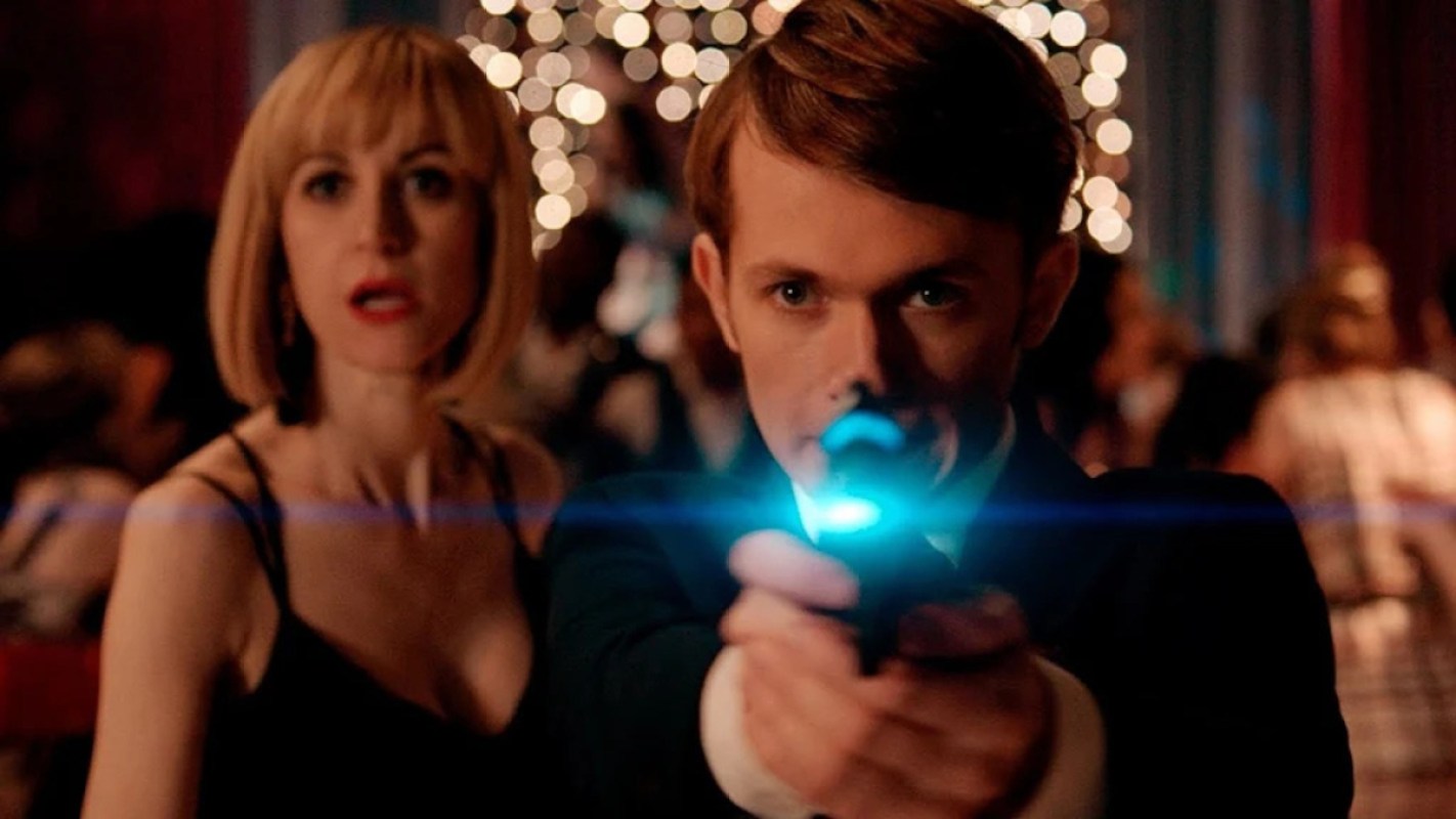 A white boy in black tie points a futuristic weapon at the camera while a blonde white woman in a black dress looks shocked behind him.