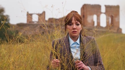 A woman in a sports coat stands in a field with ruins behind her in the distance.