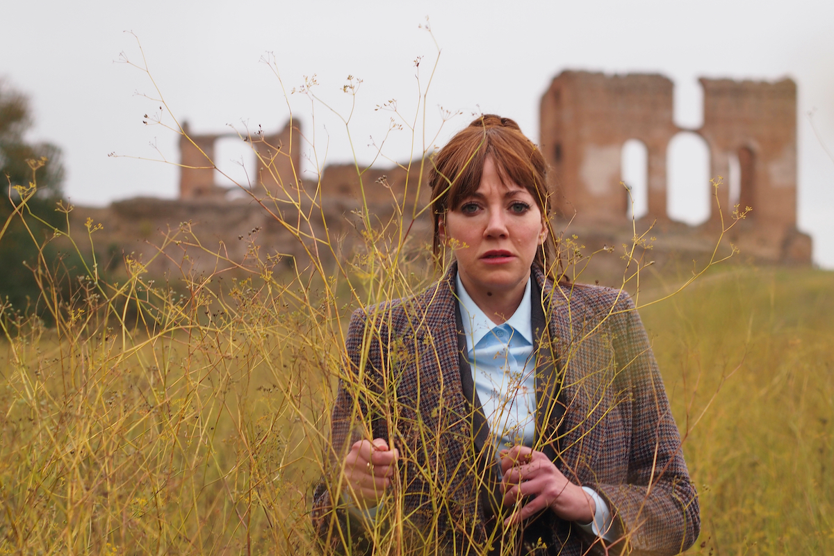 A woman in a sports coat stands in a field with ruins behind her in the distance.