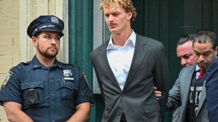 A tall white man with curly blond hair (Daniel Penny) is led out of a building by police.