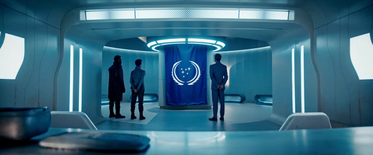 Image of David Ajala as Book, Sonequa Martin-Green as Burnham, and Adil Hussain as Sahil in a scene from the STAR TREK: DISCOVERY episode, "That Hope is You, Part 1." It's a wide shot in a minimalist, white room. From behind a table, we see Book (a Black man in a long, black coat), Burnham (a Black woman in a grey Starfleet uniform), and Sahil (a South Asian man in a grey suit) standing respectfully as they look at a blue Federation flag. 