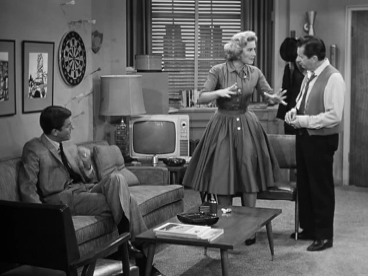 Black and white image of the fictional writers' room on the classic TV show 'The Dick Van Dyke Show.' Dick Van Dyke, a white man with short, dark hair in a light-colored suit is seated on a couch watching a blonde, female writer in a dress and a white, male writer with dark hair wearing a buttondown, vest, and dark pants have an animated discussion. 